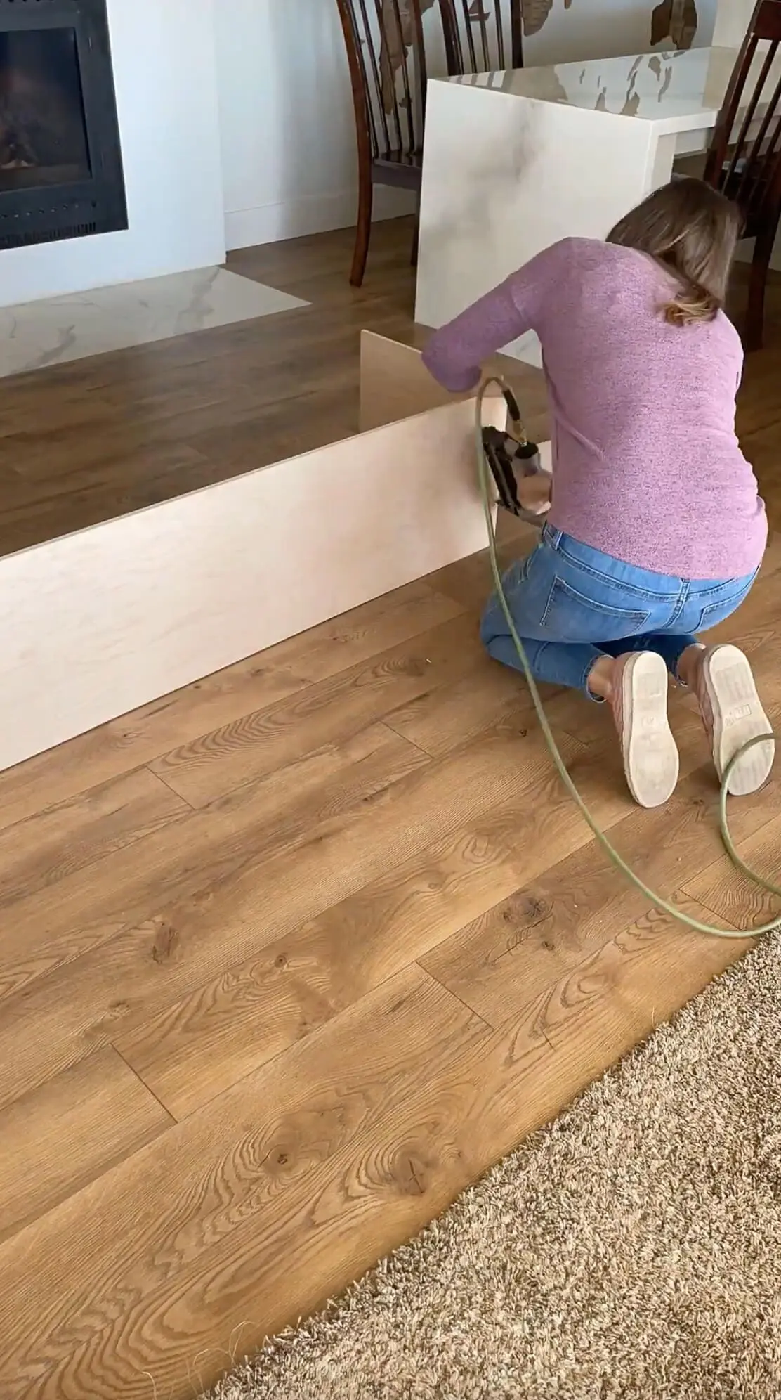 A woman using a nail gun to secure wooden panels while building a storage bench on a freshly installed wood-look floor. The space has a modern, cozy feel with a fireplace and dining area visible in the background.